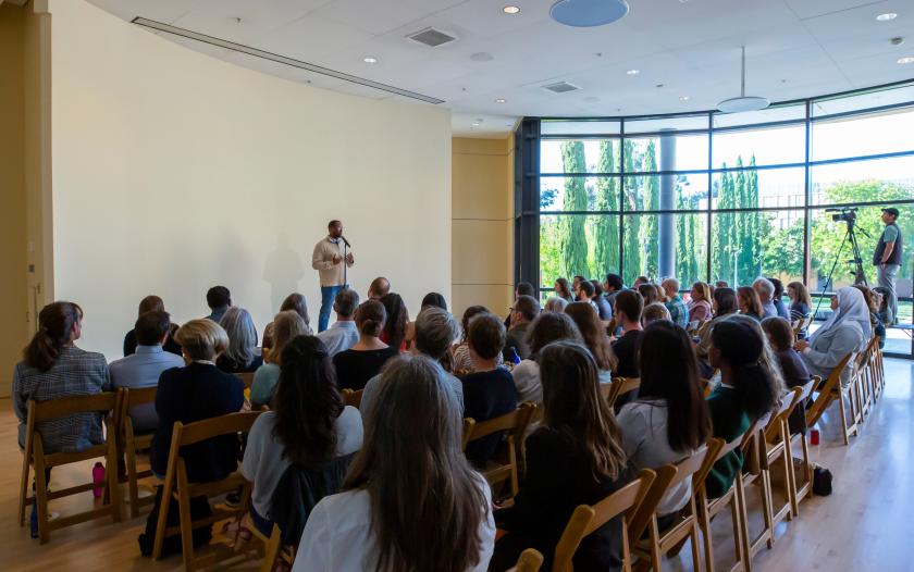 A speaker stands at a microphone in front of an audience, engaging with them. The setting is bright, with large windows showing greenery outside. The audience appears attentive, with many seated on wooden chairs. A person is visible in the background, possibly filming or monitoring the event. The atmosphere suggests an engaging presentation or discussion.