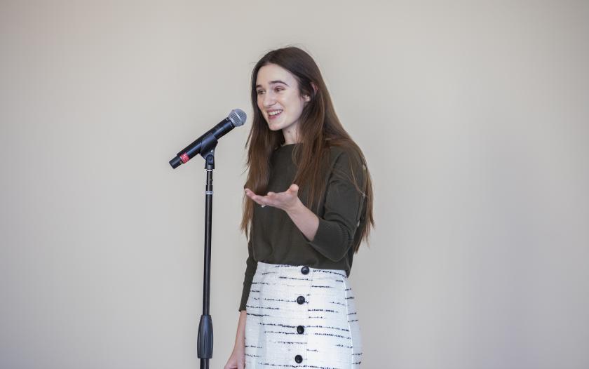 A person stands at a microphone, gesturing with one hand as they speak. They wear a dark green top and a light-colored skirt with button details. The background is plain, highlighting the speaker's expressive manner during their presentation.