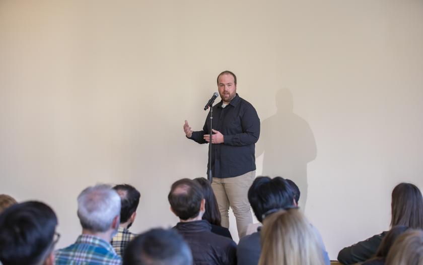 A person stands at a microphone on stage, speaking and gesturing with one hand. They wear a dark shirt and beige pants. The background is plain, and the audience, mostly turned away, is engaged in listening to the speaker.