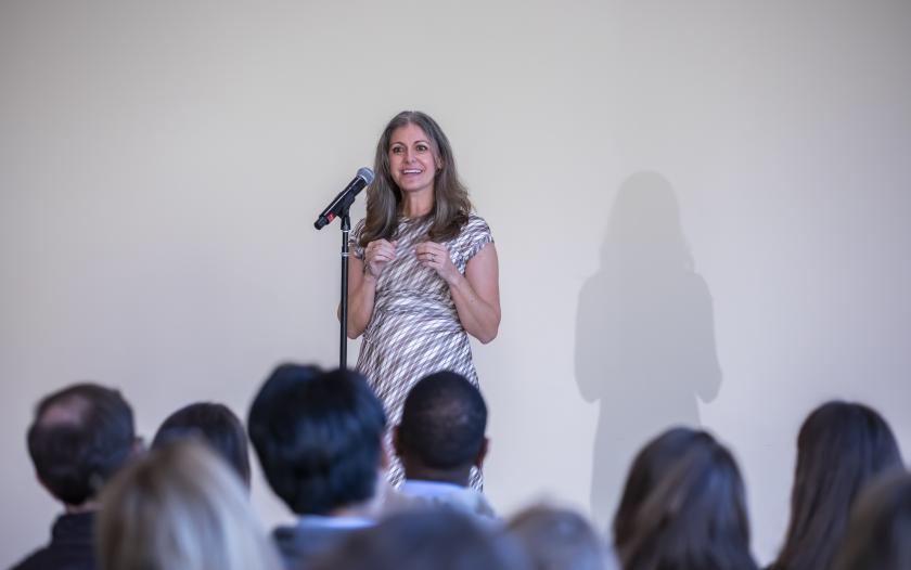 A person stands at a microphone on stage, speaking to an audience. The backdrop is plain, and the audience members, mostly turned away, are not clearly visible. The speaker wears a patterned dress and gestures with her hands while addressing the crowd.